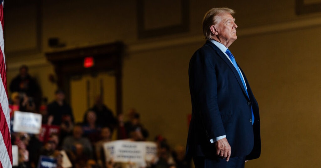 A political figure standing on stage in a dark suit and blue tie, with an American flag in the foreground and a crowd holding signs in the background, symbolizing a political rally or event.