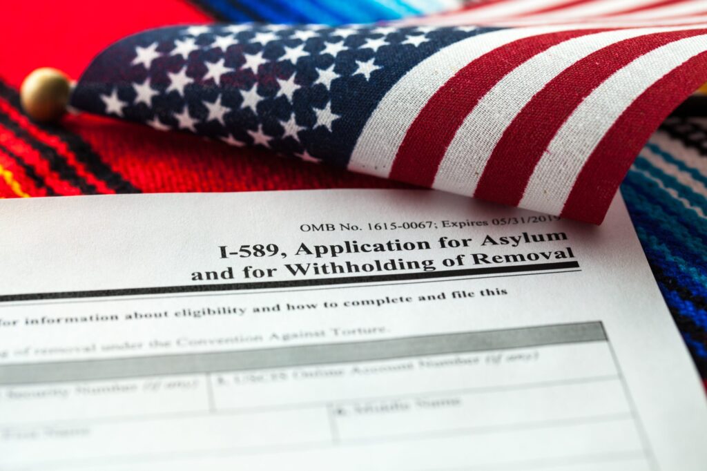U.S. I-589 Application for Asylum and for Withholding of Removal form placed on a colorful woven fabric, with a partially folded American flag in the background, symbolizing immigration and asylum seeking in the United States.