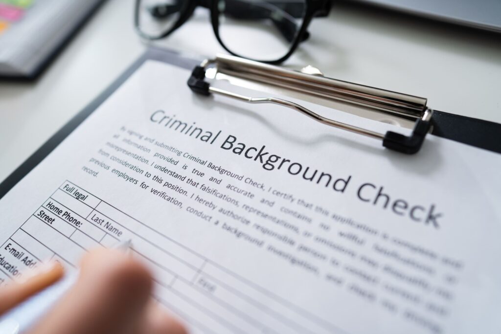 Close-up of a criminal background check form on a clipboard, with a person filling it out and a pair of glasses resting on the desk, symbolizing employment screening, legal records, or personal verification.